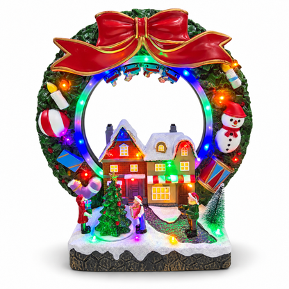 Lighted Christmas village wreath with red bow, snowman, snowy houses and miniature carolers on a white background